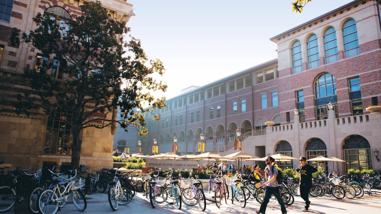 USC学生在Ronald Tutor学生活动中心前穿行,周围停放着大量自行车,阳光洒在红砖建筑与林荫小道上,充满校园氛围。