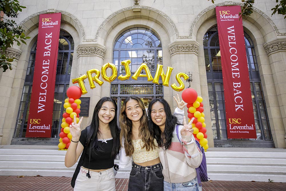 三位南加州大学商学院学生在开学日迎新活动中微笑合影,背景为Leventhal会计学院建筑和红黄气球拱门。